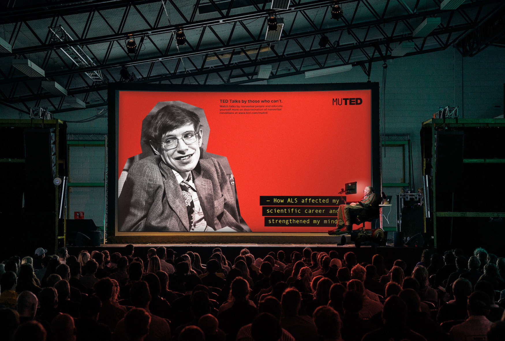 Stephen Hawking is on stage in front of a large screen, giving a talk to an audience on his non-verbal conditions.
