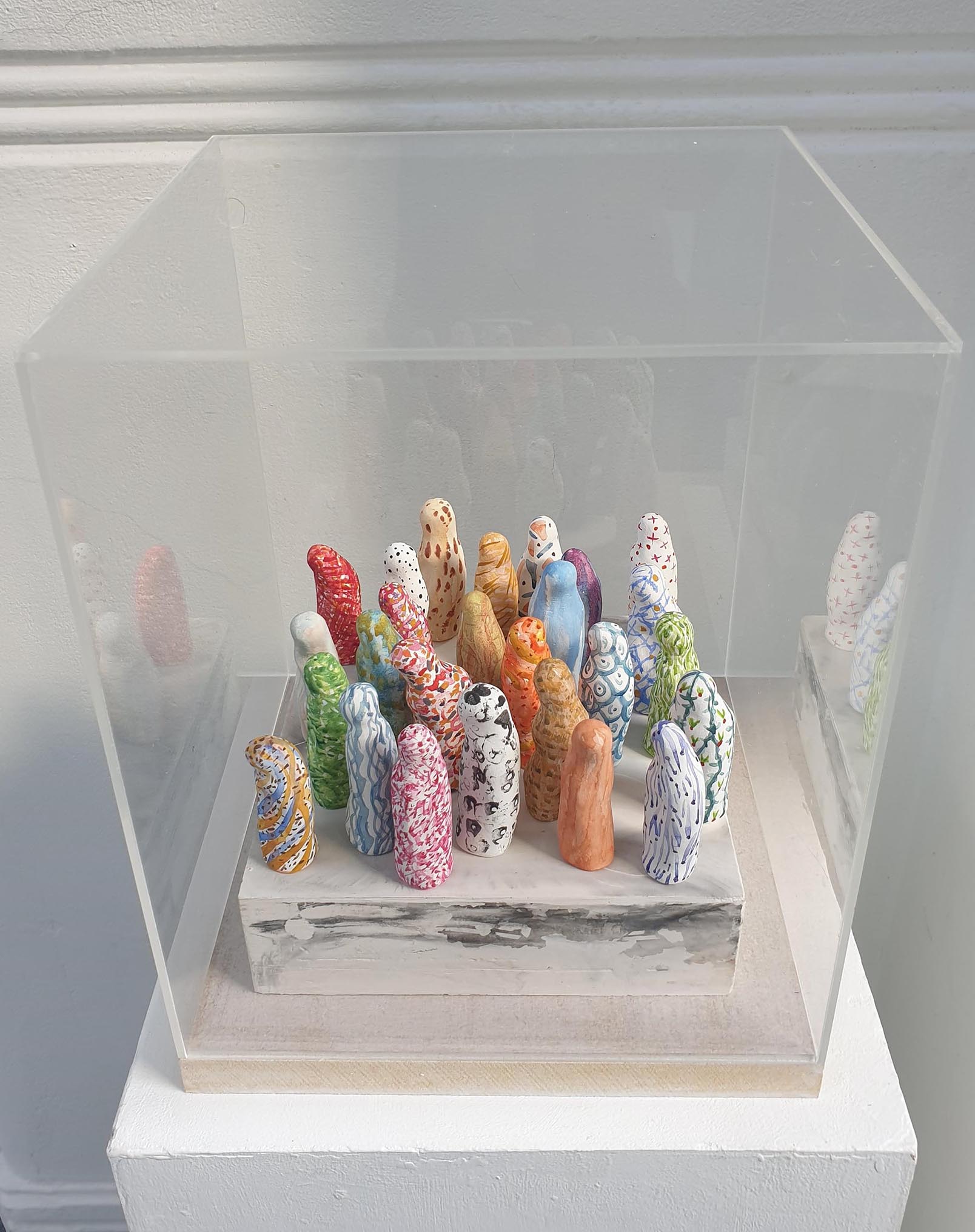 Small brightly decorated clay forms, standing close together on a marbled plaster plinth, enclosed in a transparent acrylic plastic box on a wooden base.