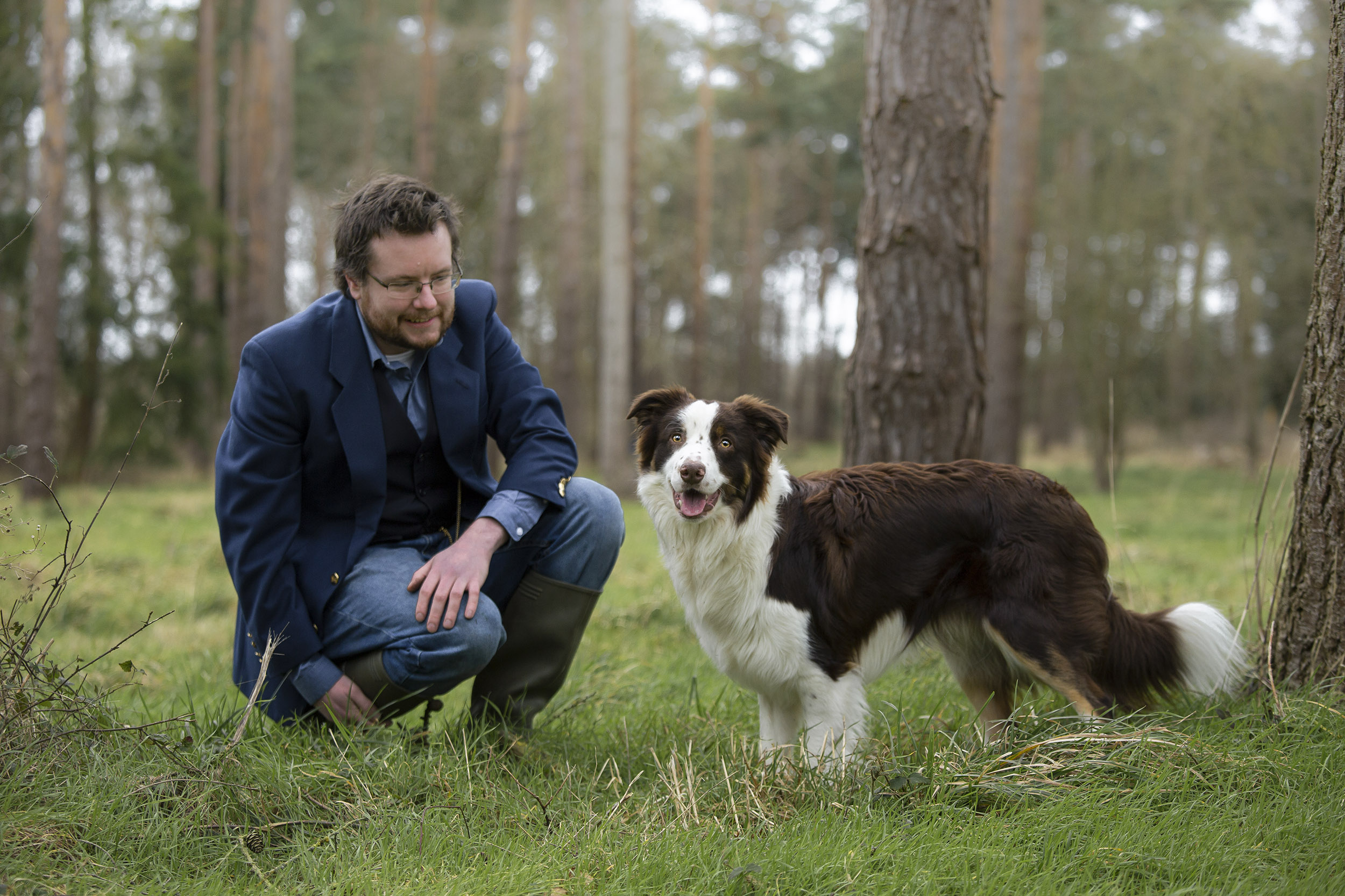 Photograph by Charlotte Hutchins showing a border collie dog and human crouched on the forest floor, looking at the camera.
