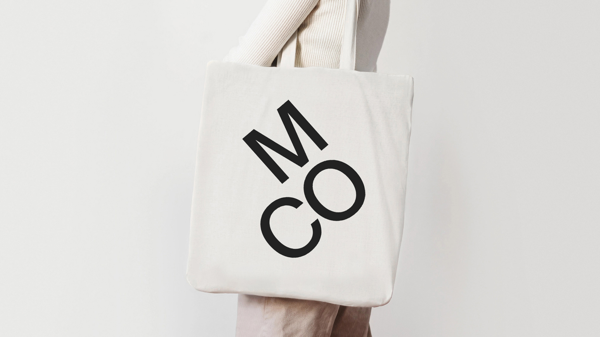 BA Graphic Communication identity by Chrysangelos Kougioutas for Museum of Communication. A model photographed from the shoulder down, dressed in neutral coloured clothing, with a tote bag over their shoulder. On the tote, the letters MOC are printed forming a right angle shape, in black sans serif typography.