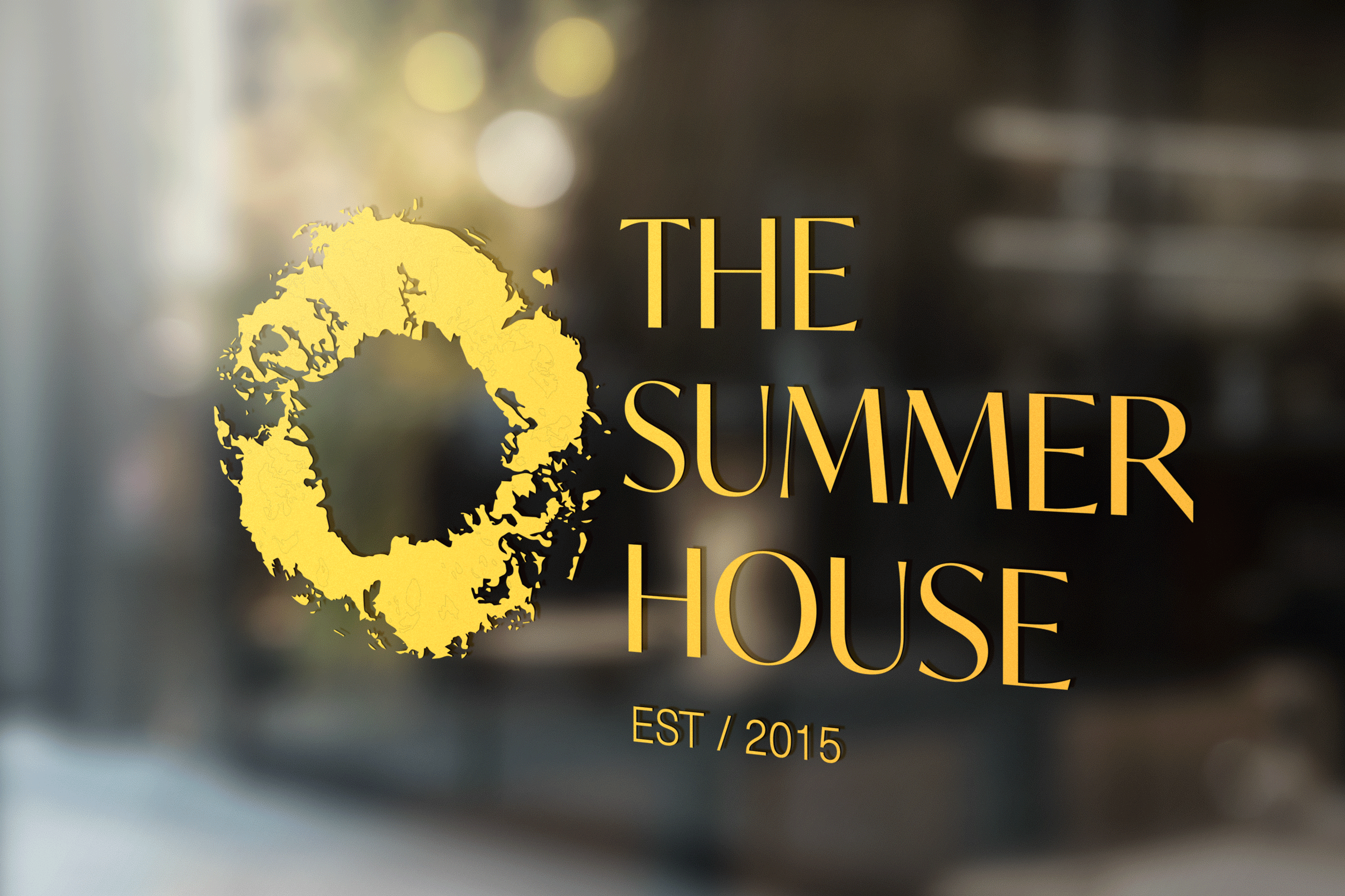 An icon and typographic logo with print made from a ring of pineapple on the left, and narrow art deco style serif type which reads THE SUMMER HOUSE, all in gold foil on a glass surface