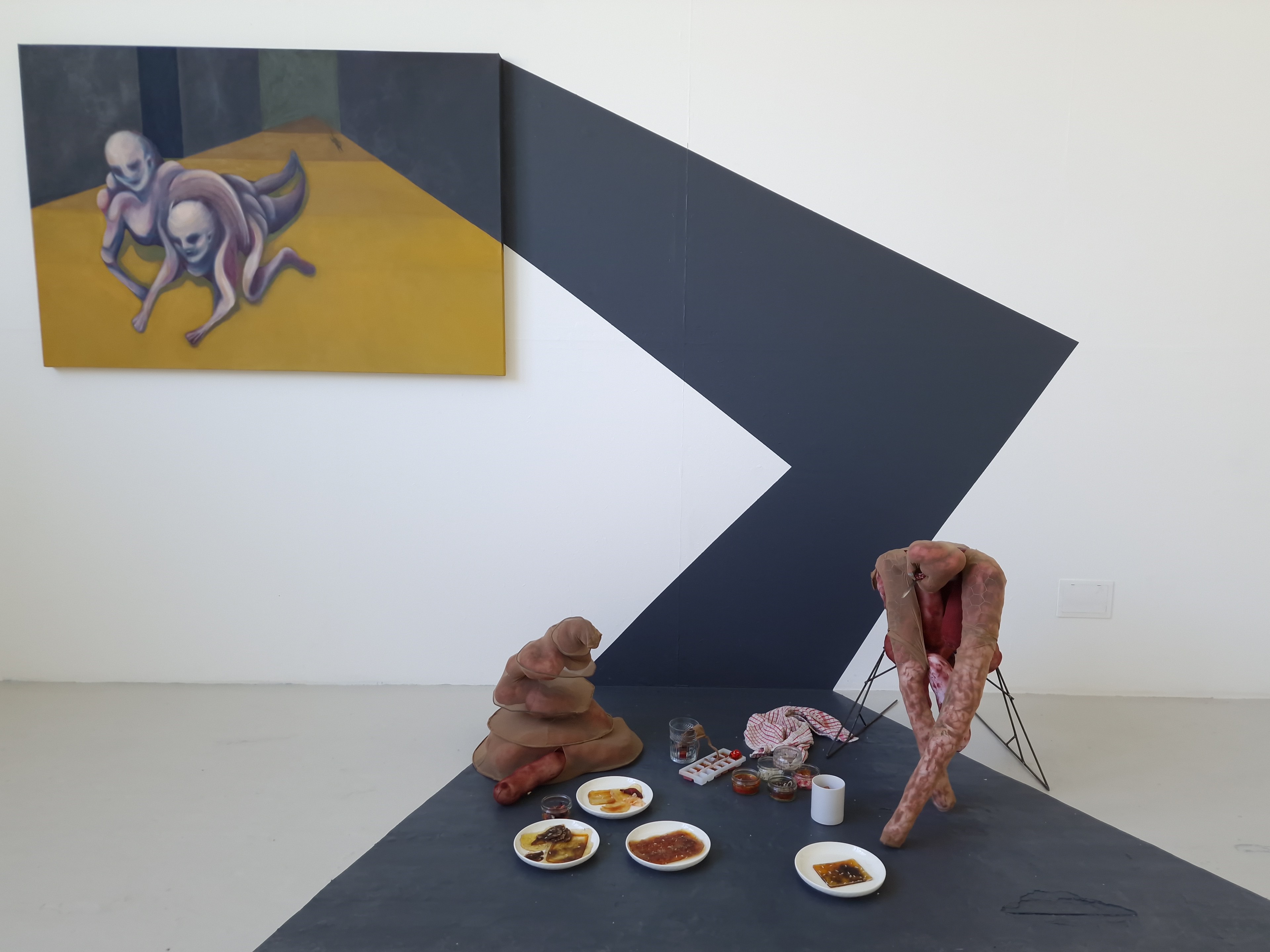 An installation by Elena Genovese depicts a painting of two figures and an animal travelling through a corridor and multiple sculptures on the floor. A grey stripe connects the painting and the picnic.
