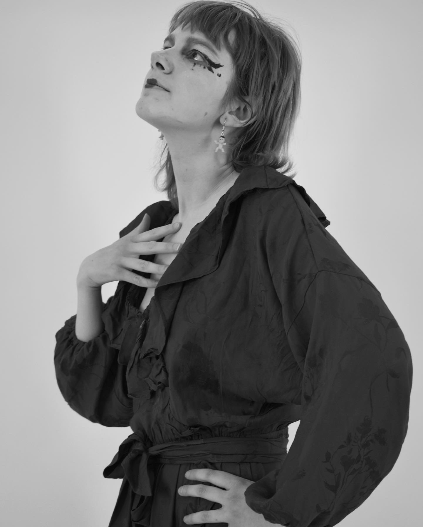 A monochrome image of a woman facing the left, looking upwards. One hand on her chest, one on her hip. She wears bold eyeliner, a thrilled long sleeved dress and earrings that are babies.