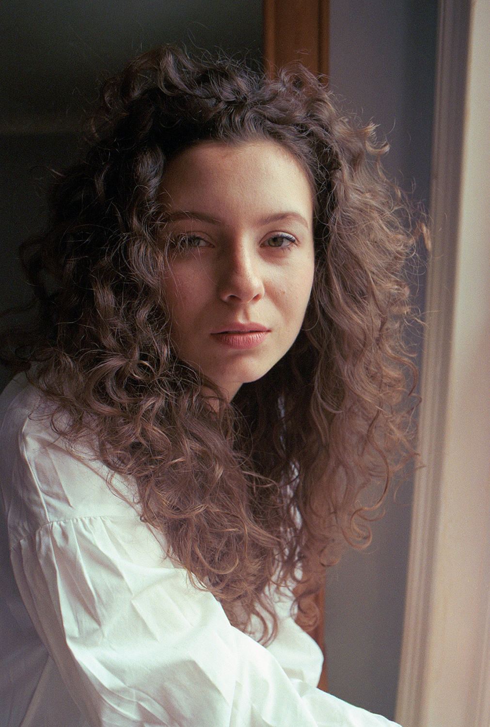 Woman standing by her window, staring into the camera, she is wearing a white shirt and has big curly hair.