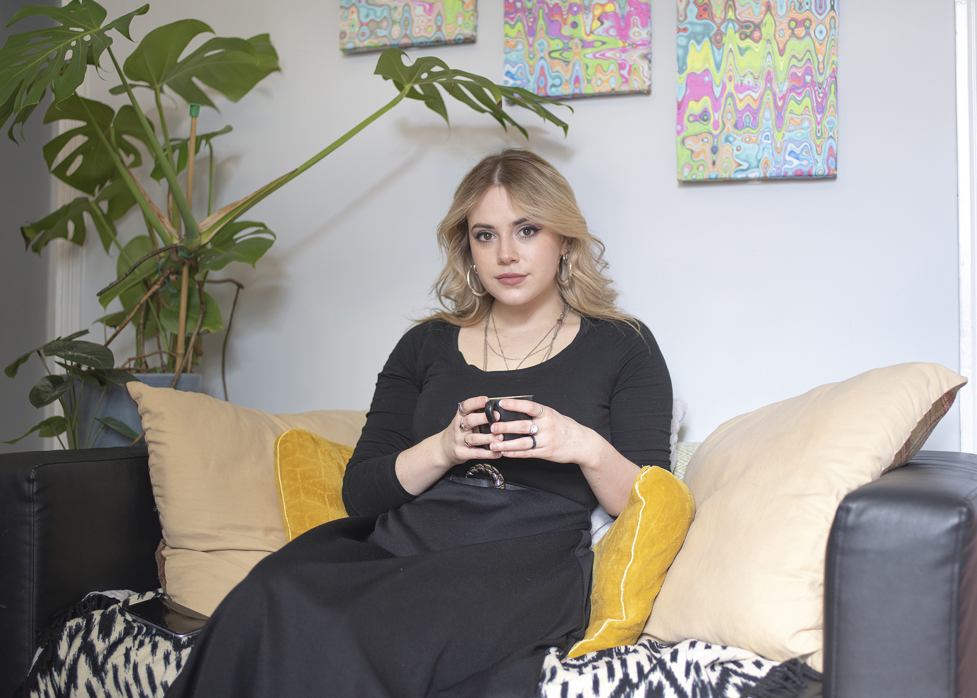 BA Photography work by Hannah Jayne showing a documentary style portrait of a blonde woman seated in her living room surrounded by plants and artwork.