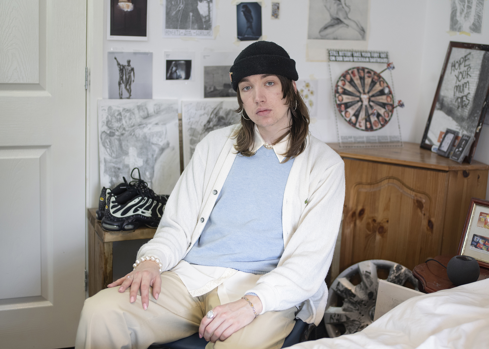 BA Photography work by Hannah Jayne showing a documentary style portrait of a male dressed in cream tones, sat in his room surrounded by prints and art work plastering his walls.