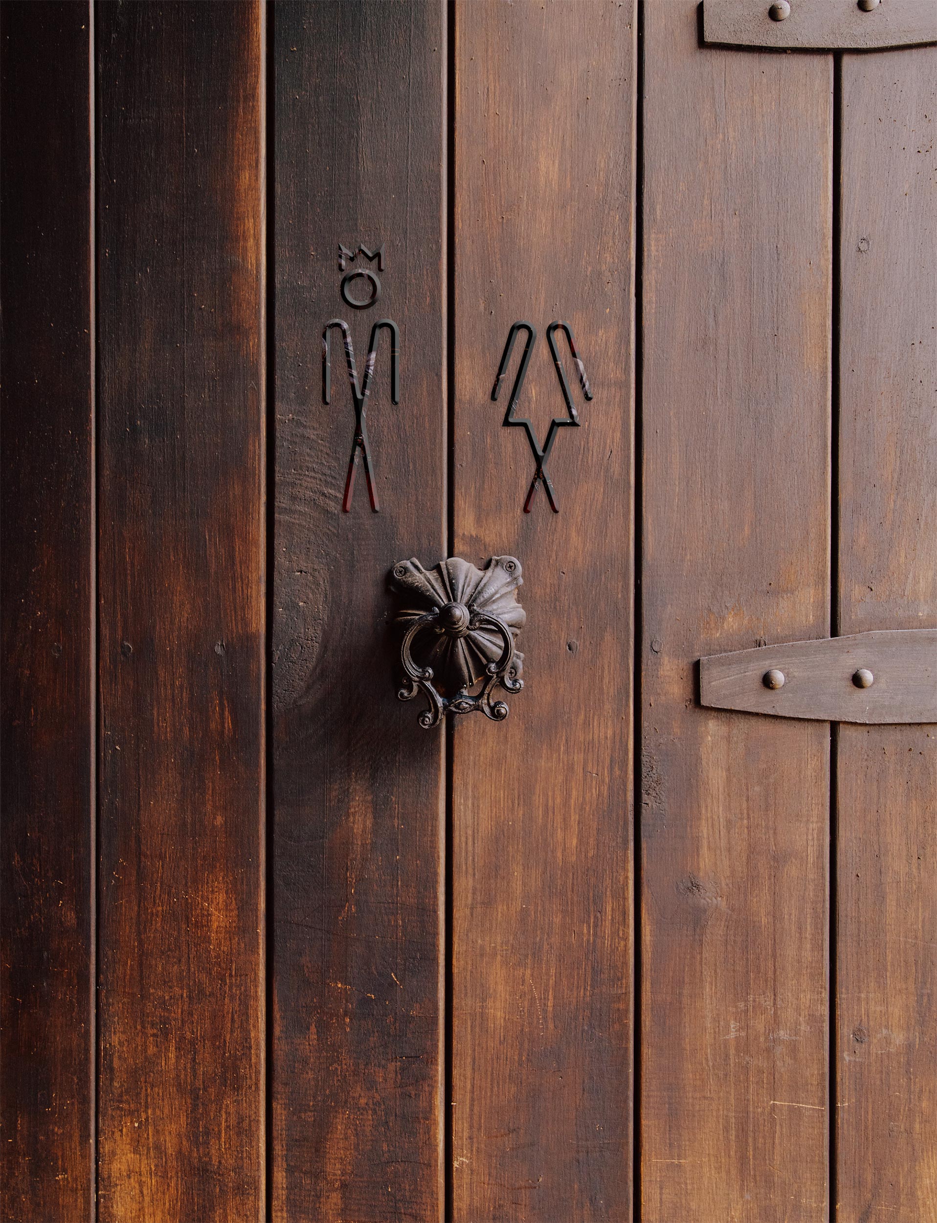 Photo of a wood panelled toilet door, with a knocker in the centre. Above, wire outlines of male/female symbols for toilets. The male figure has a crown, representing Henry VIII, and the female figure has lost her head, representing Anne Boleyn.