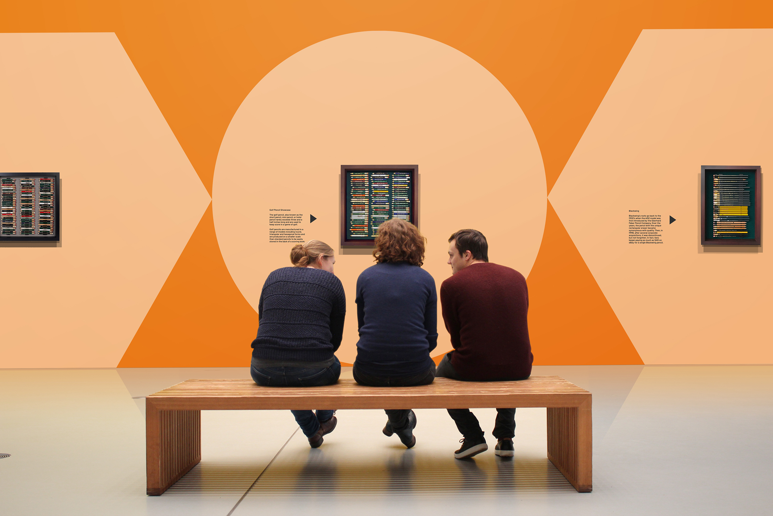 Graphic Communication work by James Keeley showing Derwent Pencil Museum Environment. Three people sit on a bench in the centre of an empty gallery. The wall in front of them is bright orange, with large pale orange graphic shapes of pencils.