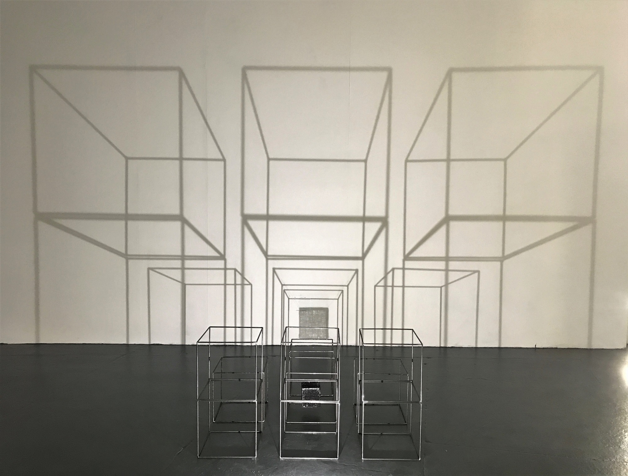 Sculpture by Jo Bellamy. Steel cubes and a smaller fragile thread cube, stacked and lit creating dramatic large shadows resembling large structures.