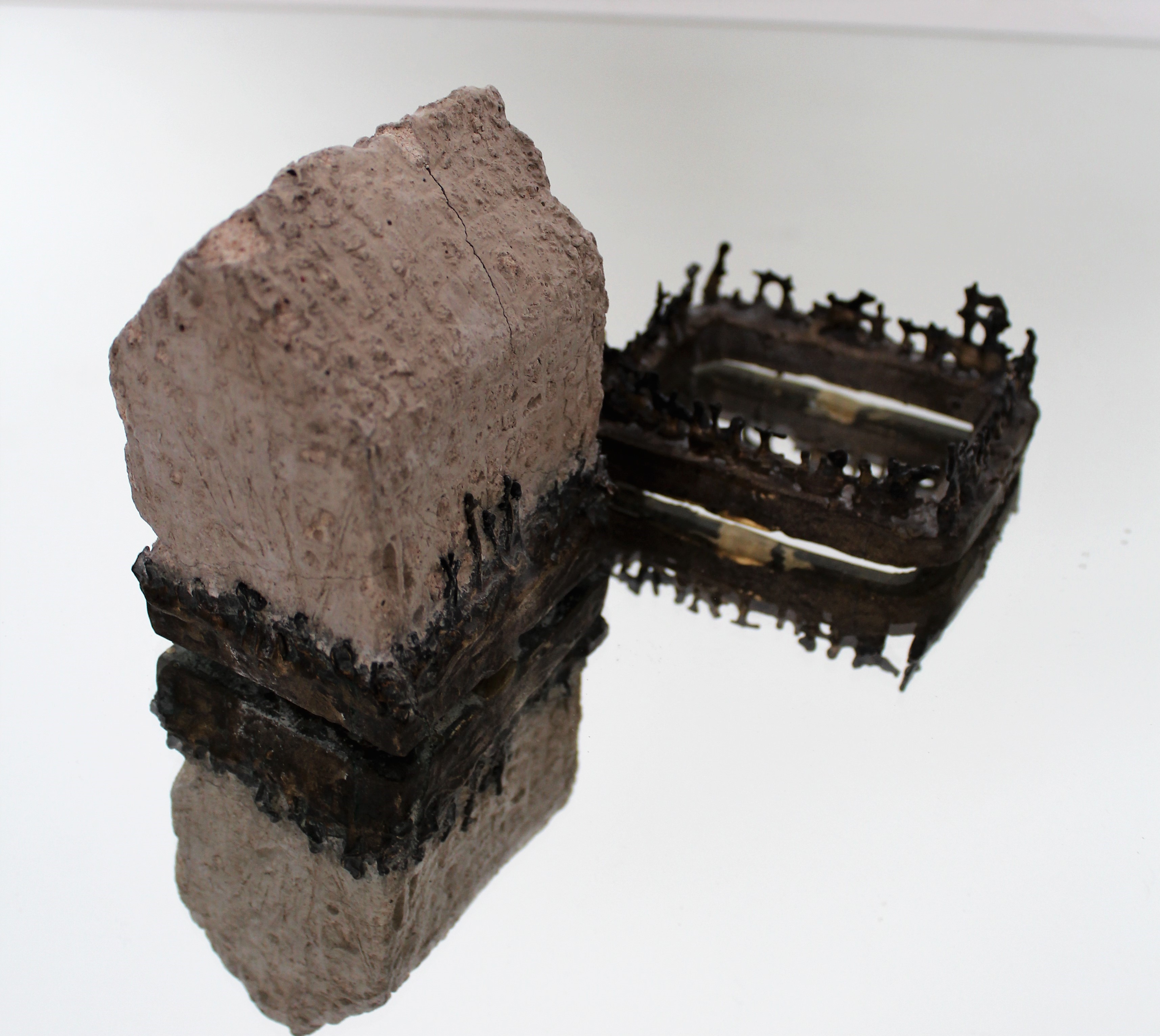 Sculpture by Jo Bellamy. Two small bronze forms, one with a plaster house form attached, the second only a base of a similar size, with the remnants of mesh sides visible. Placed on a mirrored base.