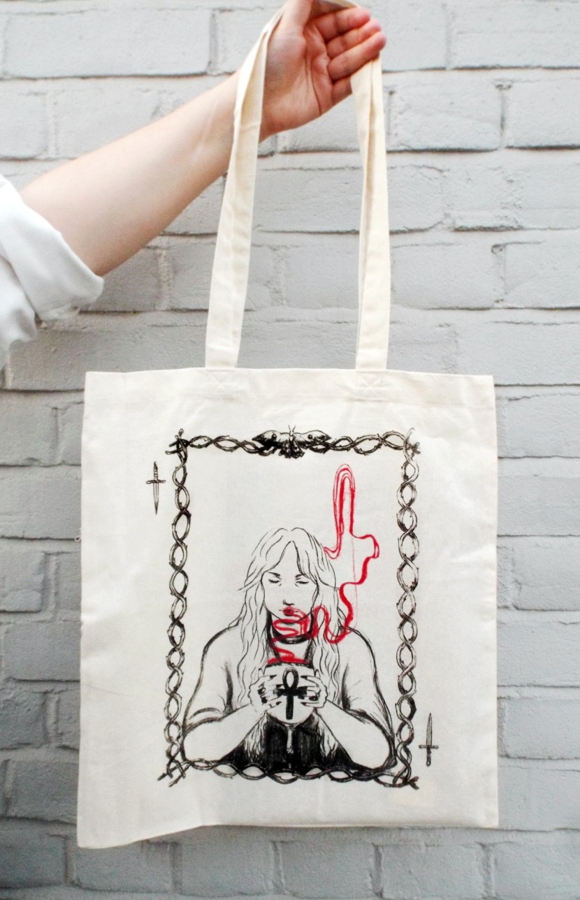 An image of a hand holding up a cream tote bag, which has an illustration of a witch consuming eternal life, by BA Illustration graduate Leah McGhee