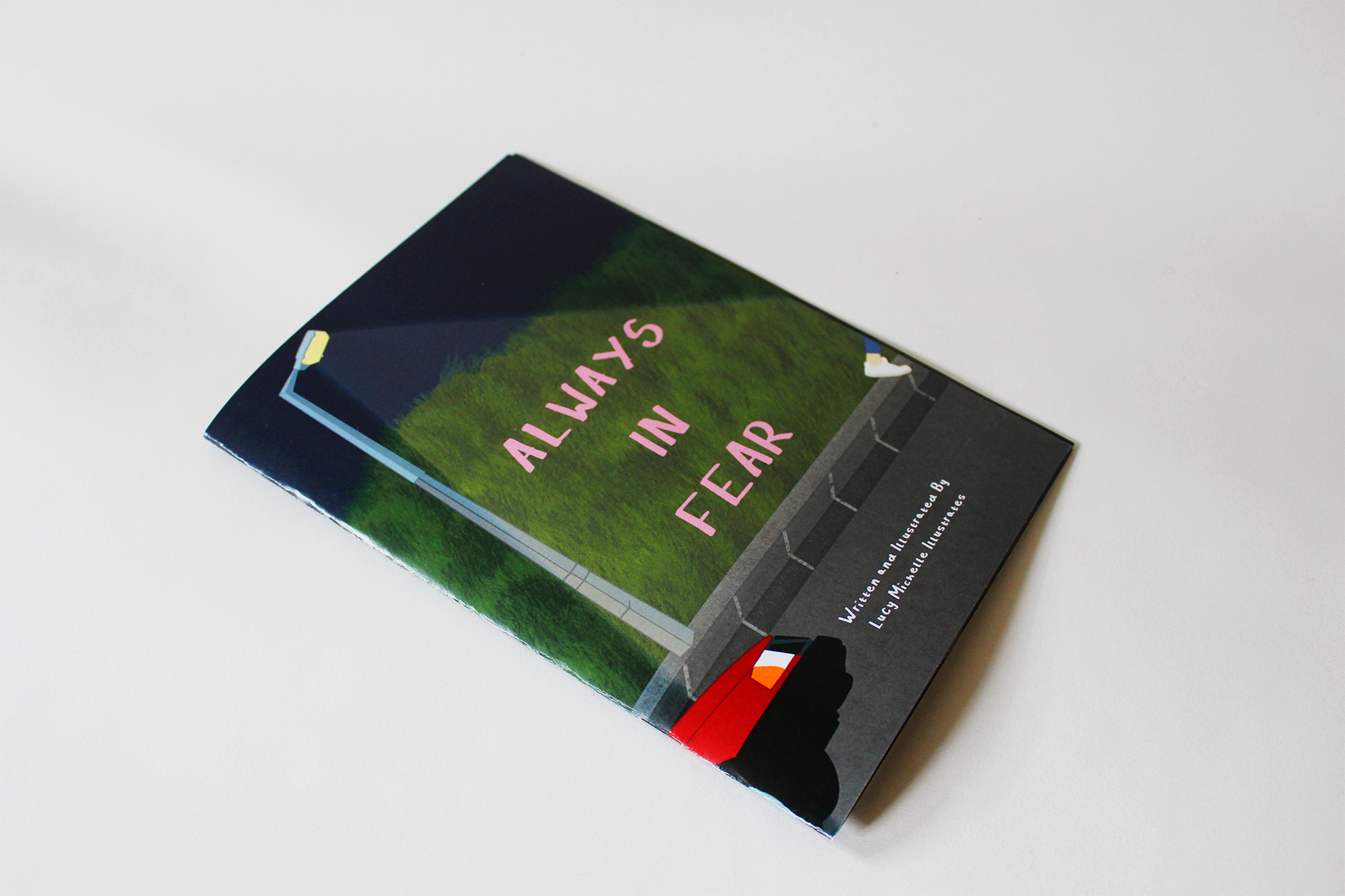 A photograph of printed zine 'Always in Fear' by Lucy Taylor. The front cover shows a road and pathway, lined by green hedges. Creeping on to the side of the zine is a red car, with headlights on. A lamppost shines a light downward, revealing the title of the zine. A small foot can be seen leaving the other side of the page, as if walking out of the frame