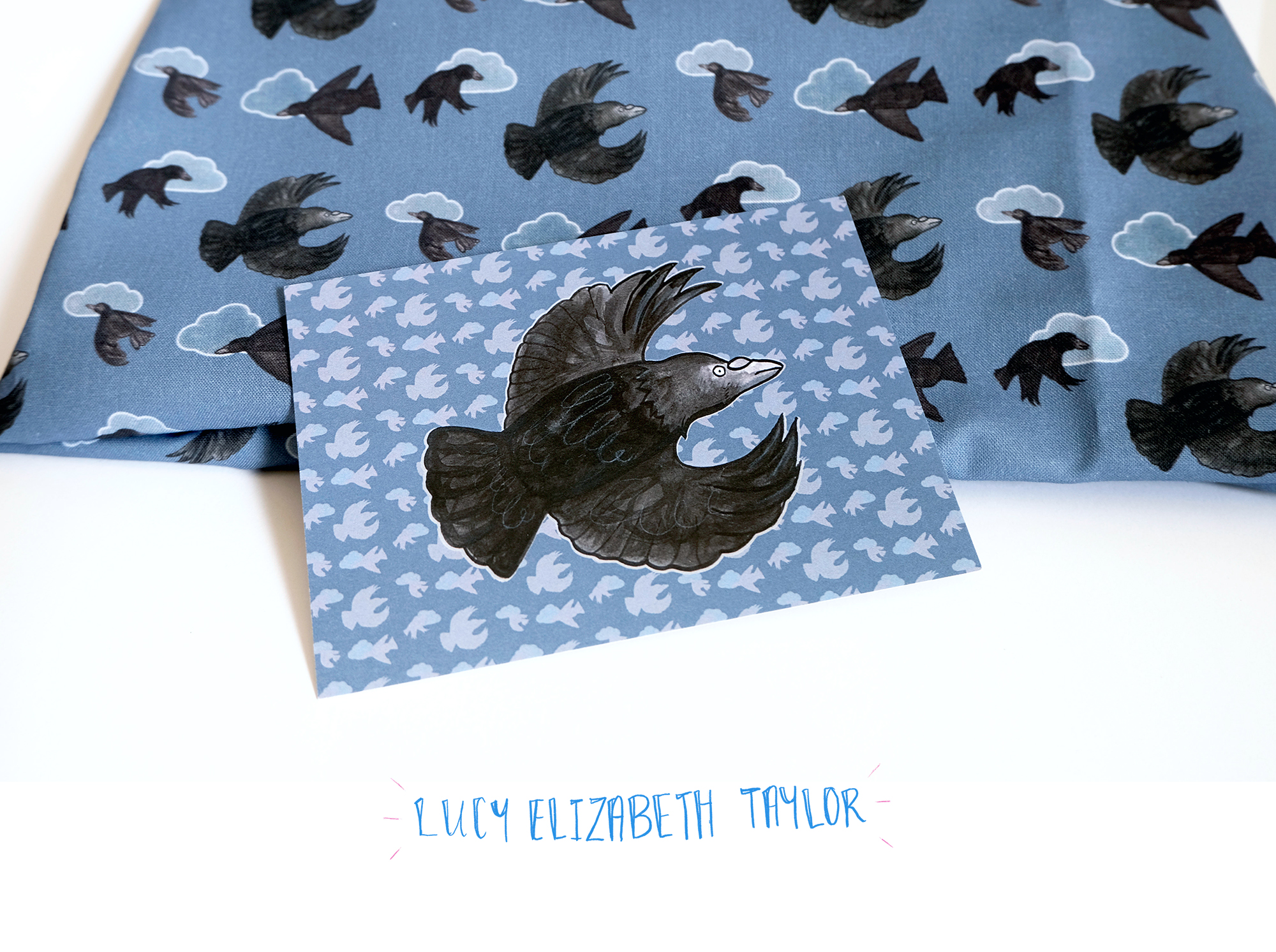 A folded piece of fabric featuring crow and cloud pattern, rested on top is a postcard also featuring Lucy Elizabeth Taylor's flying crow design.