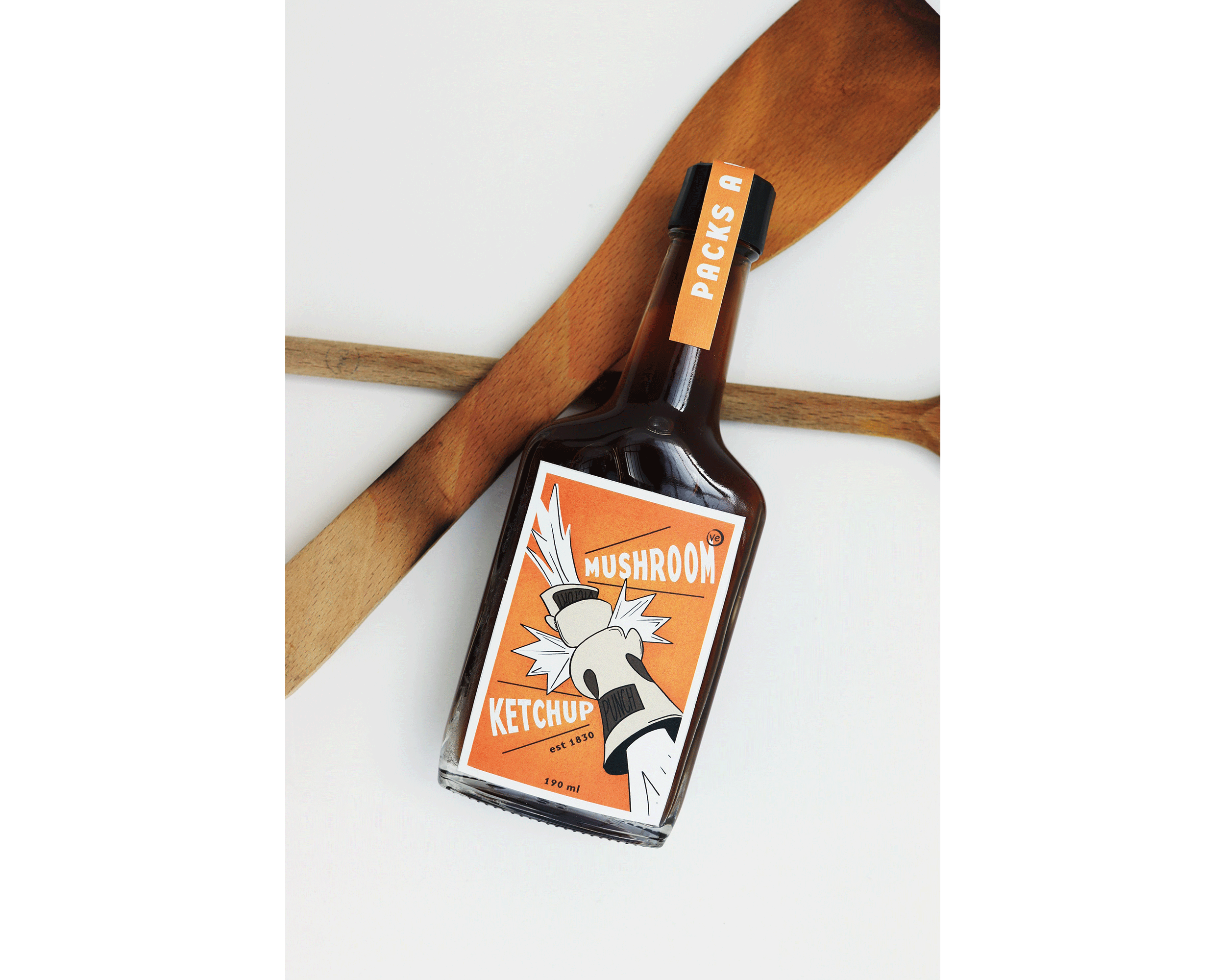 Graphic Design Packaging by Maika Elsley Medina showing a bottle laying on kitchen utensils, with a bright orange label showing a mushroom cross boxing glove punching into the design.