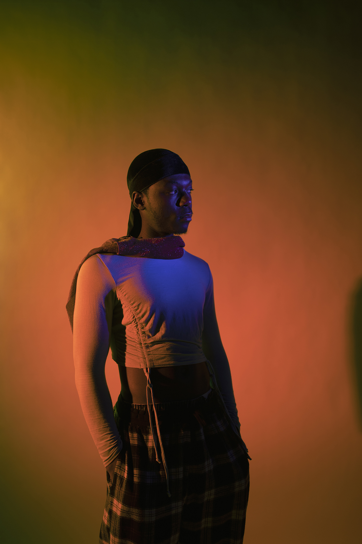 A portrait of a black man in front of an orange background. He wears a light blue crop top, checkered trousers, and a black durag.