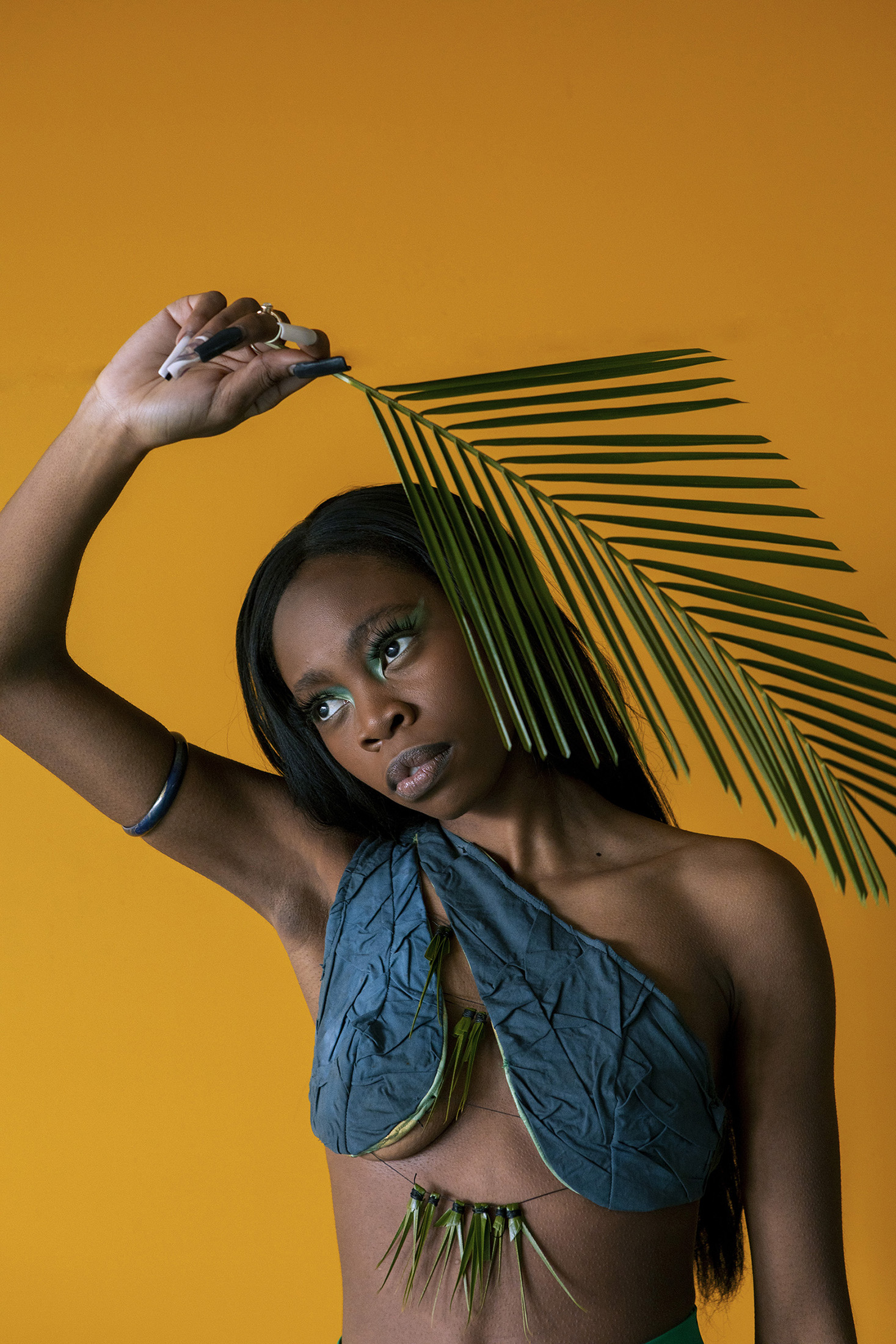 A portrait of a black woman in a handmade bralette. She stands in front of a vibrant yellow carpark wall, holding a large green leaf over her head.