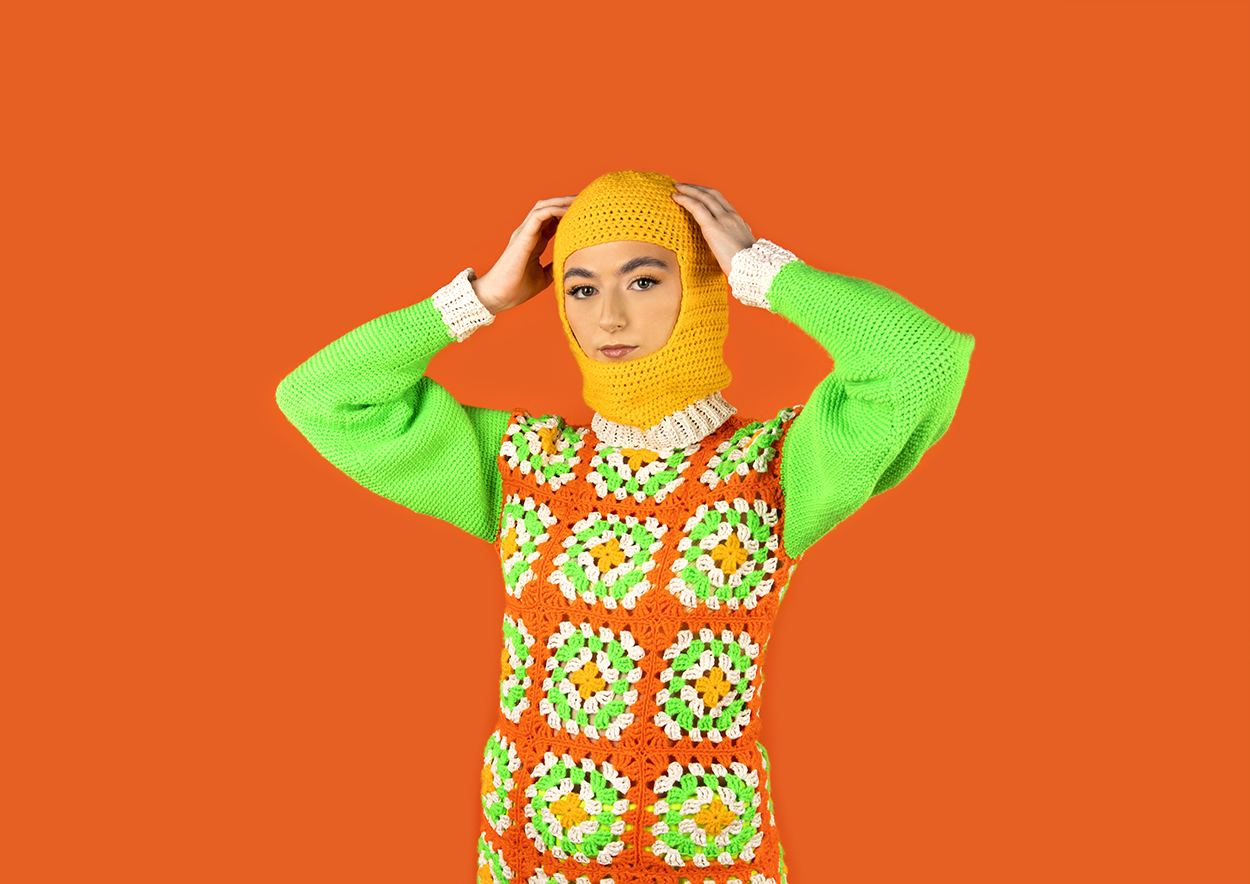 BA Photography work by Becca Stevenson showing a girl in a colourful, crochet jumper and balaclava against an orange backdrop.