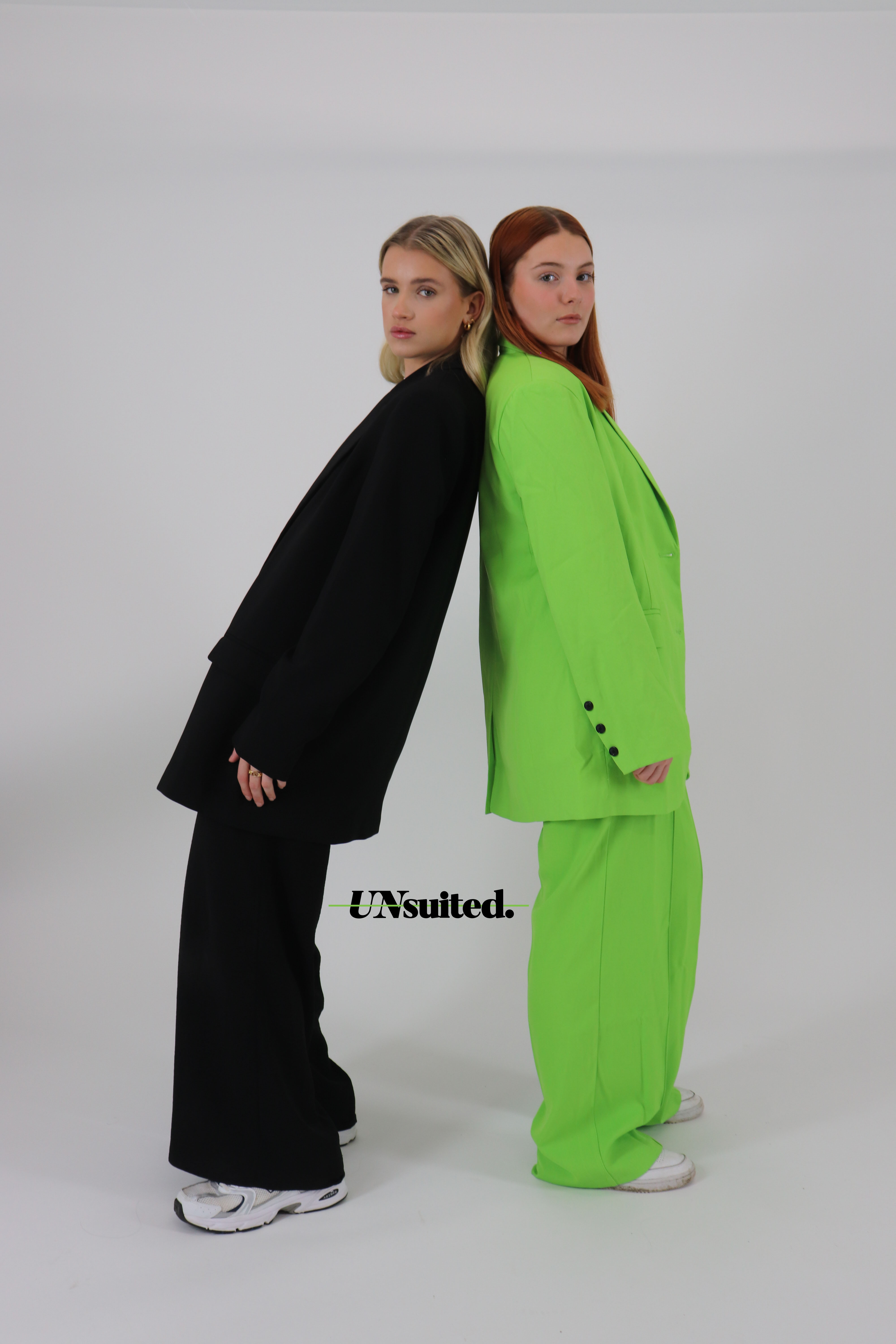 Two models in baggy suits and white trainer lean back to back. The left wears black, the right wears lime green. The text in the middle reads 'UNsuited'