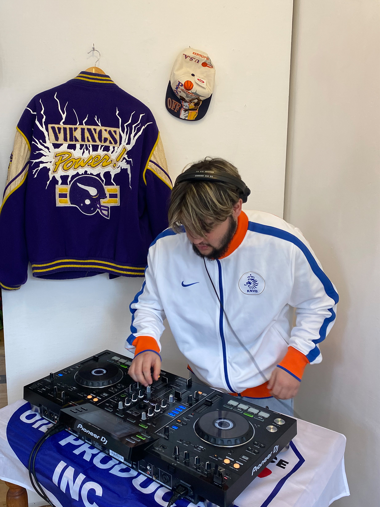 Photo of someone using a DJ deck in the corner of a room, wearing sportwear and with sportswear hung on the wall behind