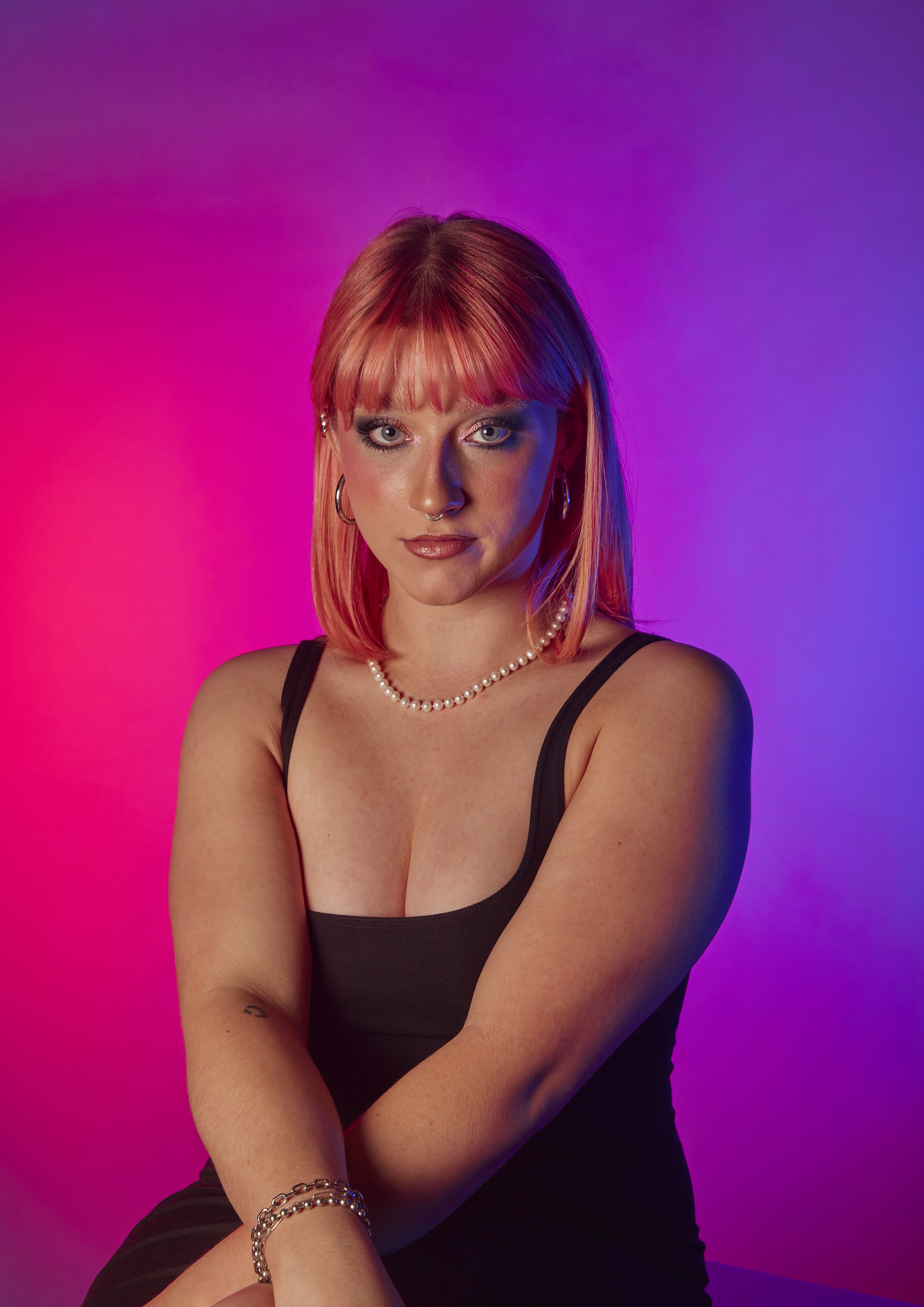 Portrait photo of model with pink hair and bangs, sitting with her arms folded in front of her. Image has a vibrant pink and purple gradient background