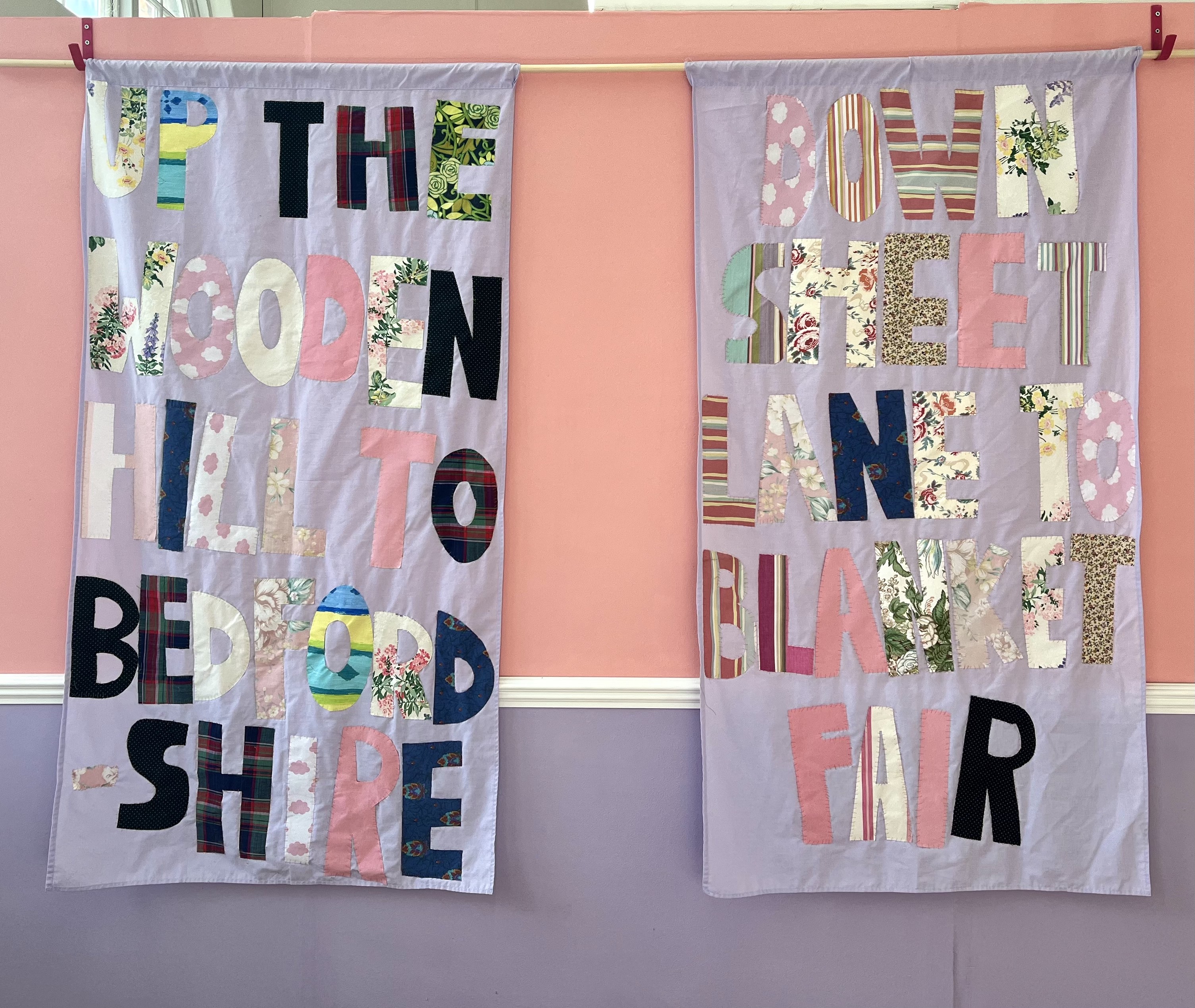 Textile hanging by Amber-Rose Thorpe, the hangings have hand-sewn applique on them that reads 'UP THE WOODEN HILL TO BEDFORDSHIRE, DOWN SHEET LANE TO BLANKET FAIR'.