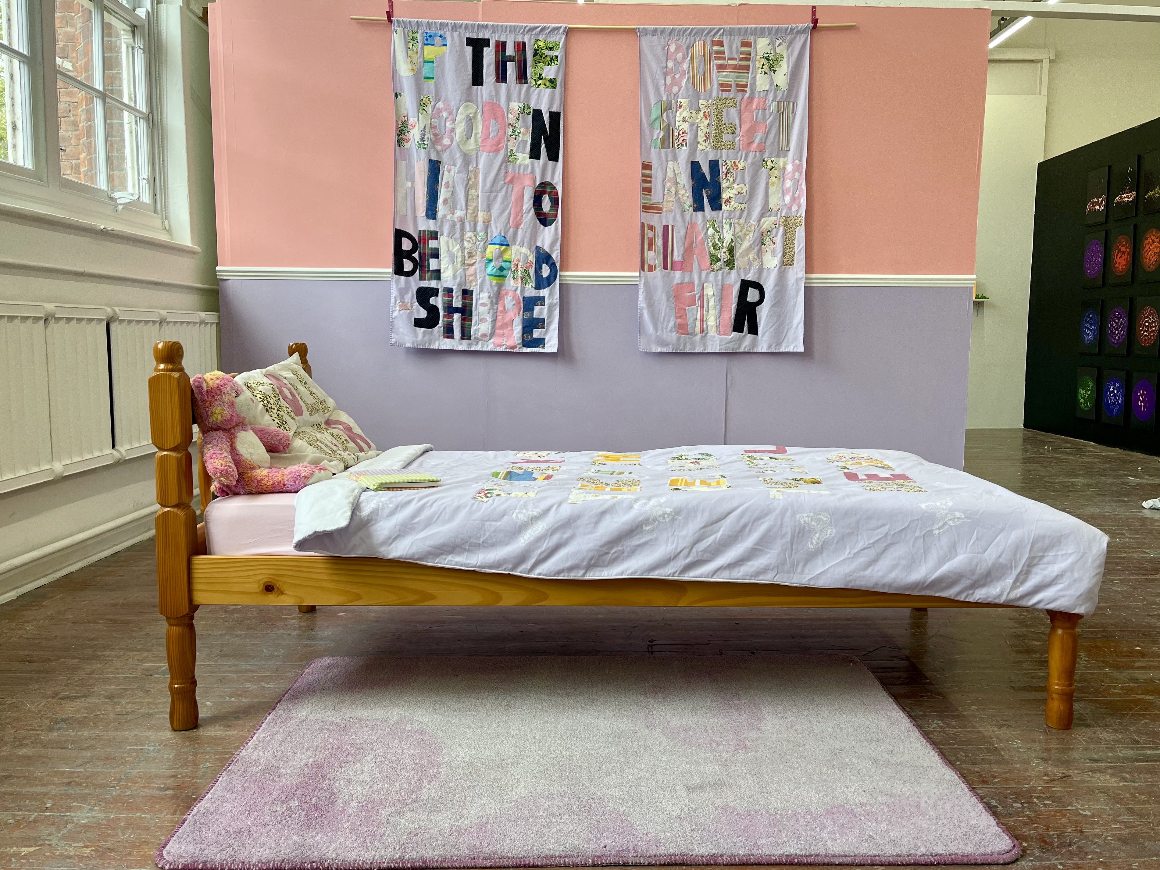 Installation by Amber-Rose Thorpe, a bed is in the foreground and a pink and purple wall is in the background. There is text on textile hangings throughout.