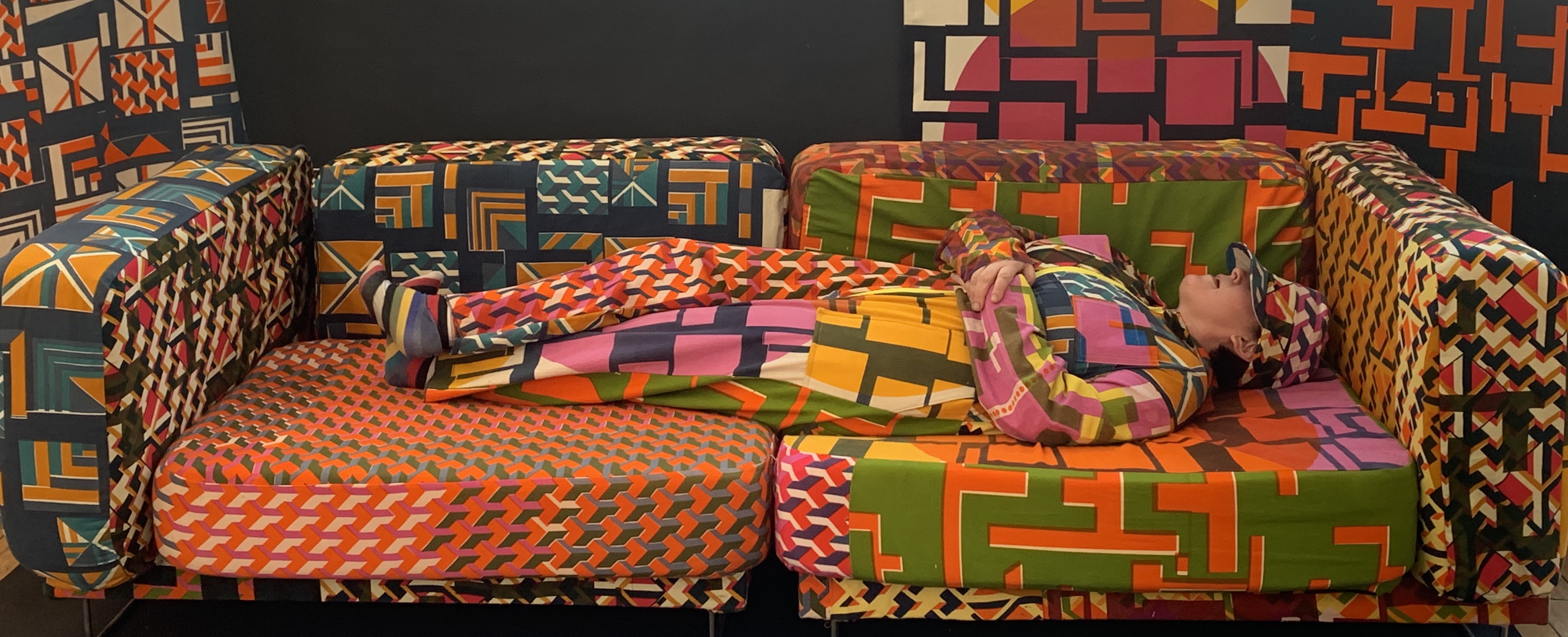 Model in boilersuit lies on sofa. The colourful array of screen-printed patterns are designed to work together on the sofa and boilersuit to camouflage the model.