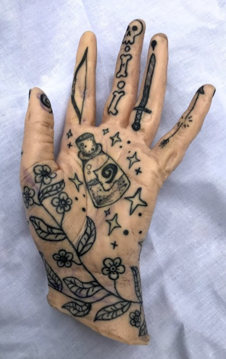 White flesh coloured silicone hand covered in black tattoos. Tattoo in the middle of the palm is of a potion bottle, there's a flower vine around the wrist and up the thumb. On the fingers are a bow, a skull and bones, a sword, and a broken arrow.