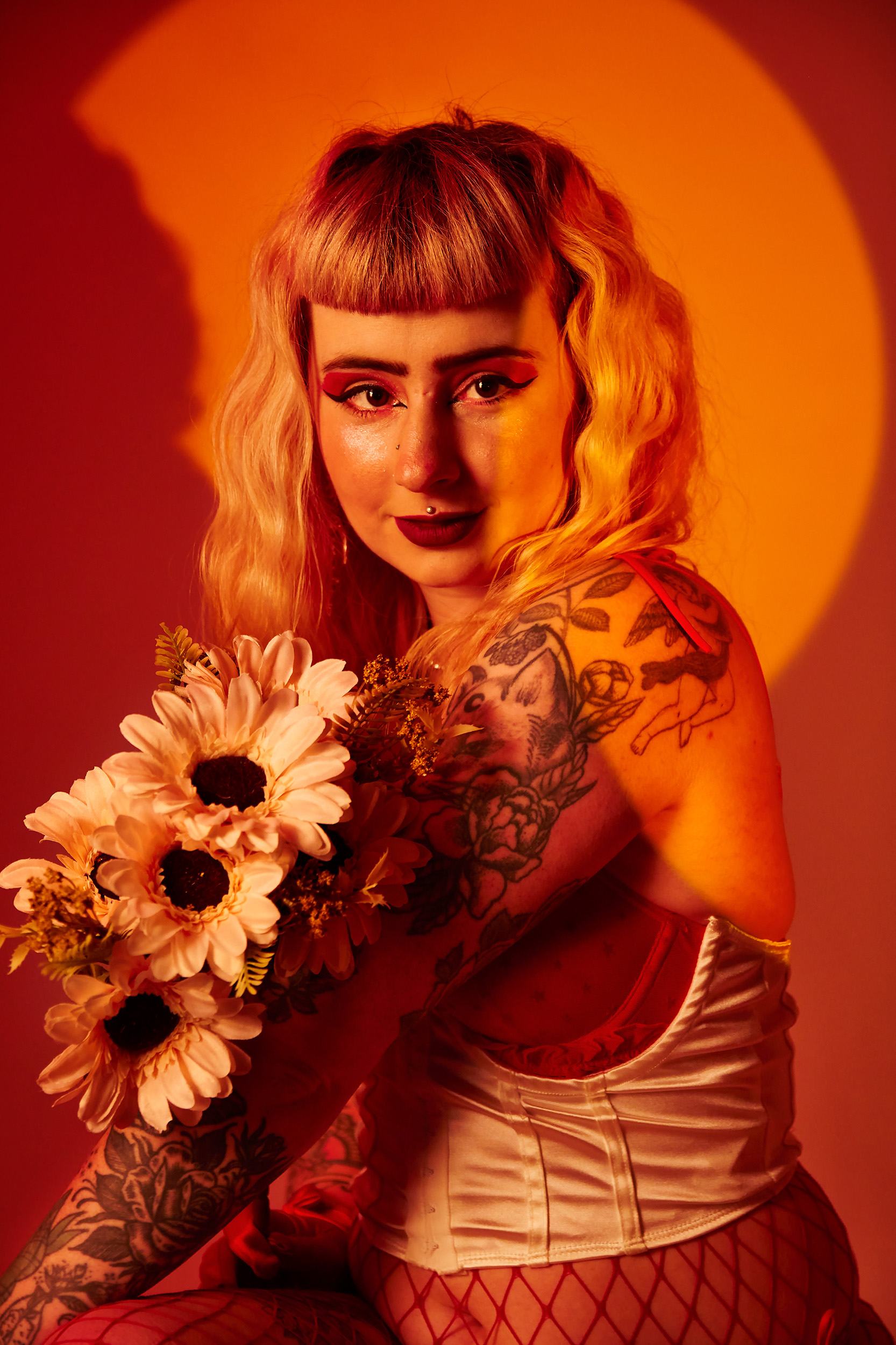 Portrait Photography by Chloe Sibley, showing a burlesque performer holding a bunch of flowers, looking straight down the lens with an orange and red spotlight encasing her.