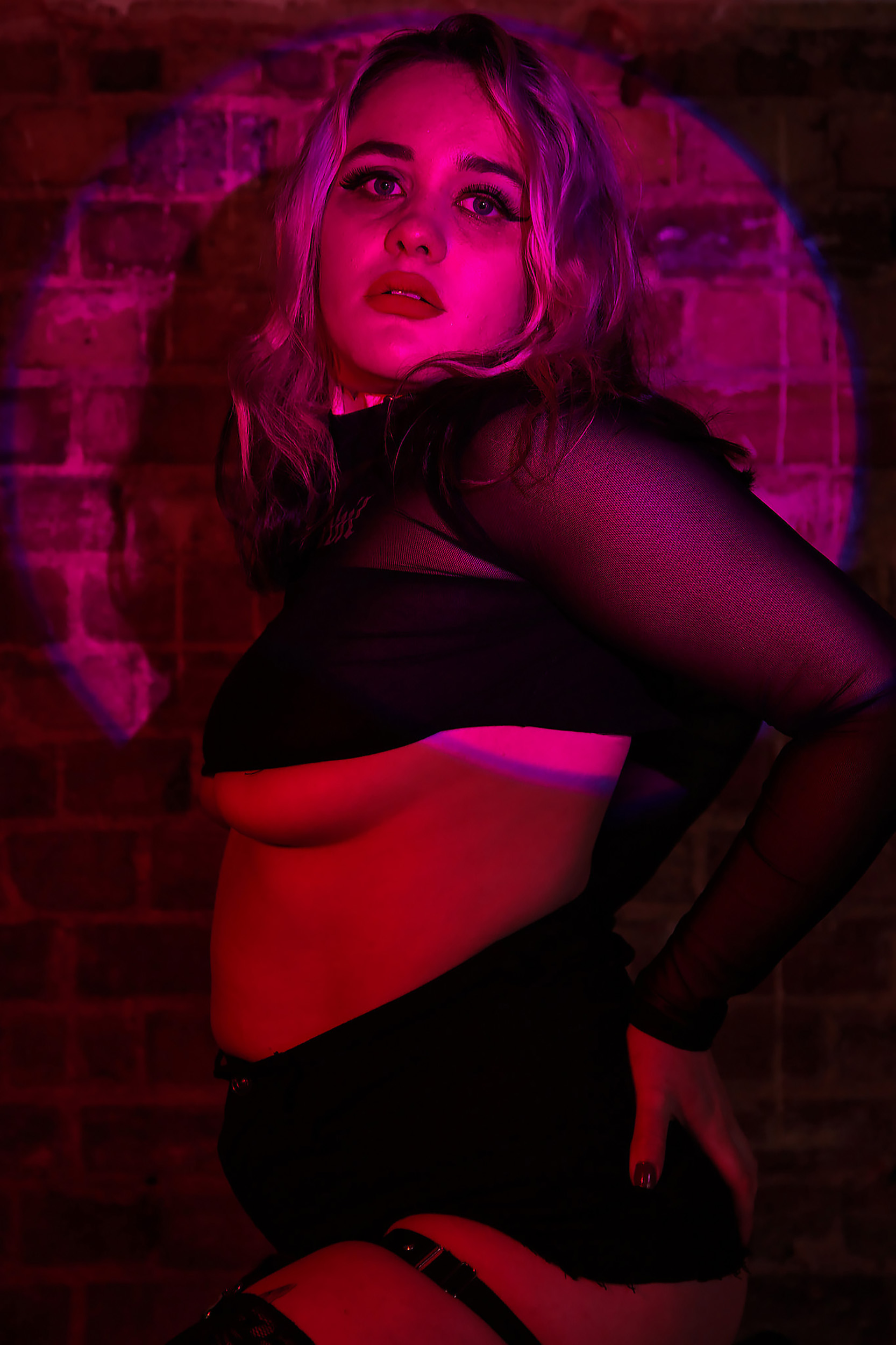 Portrait Photography by Chloe Sibley depicting a femme burlesque performer in front of a brick wall, with purple and pink spotlighting surrounding her, wearing a black crop top that leaves some underboob on display
