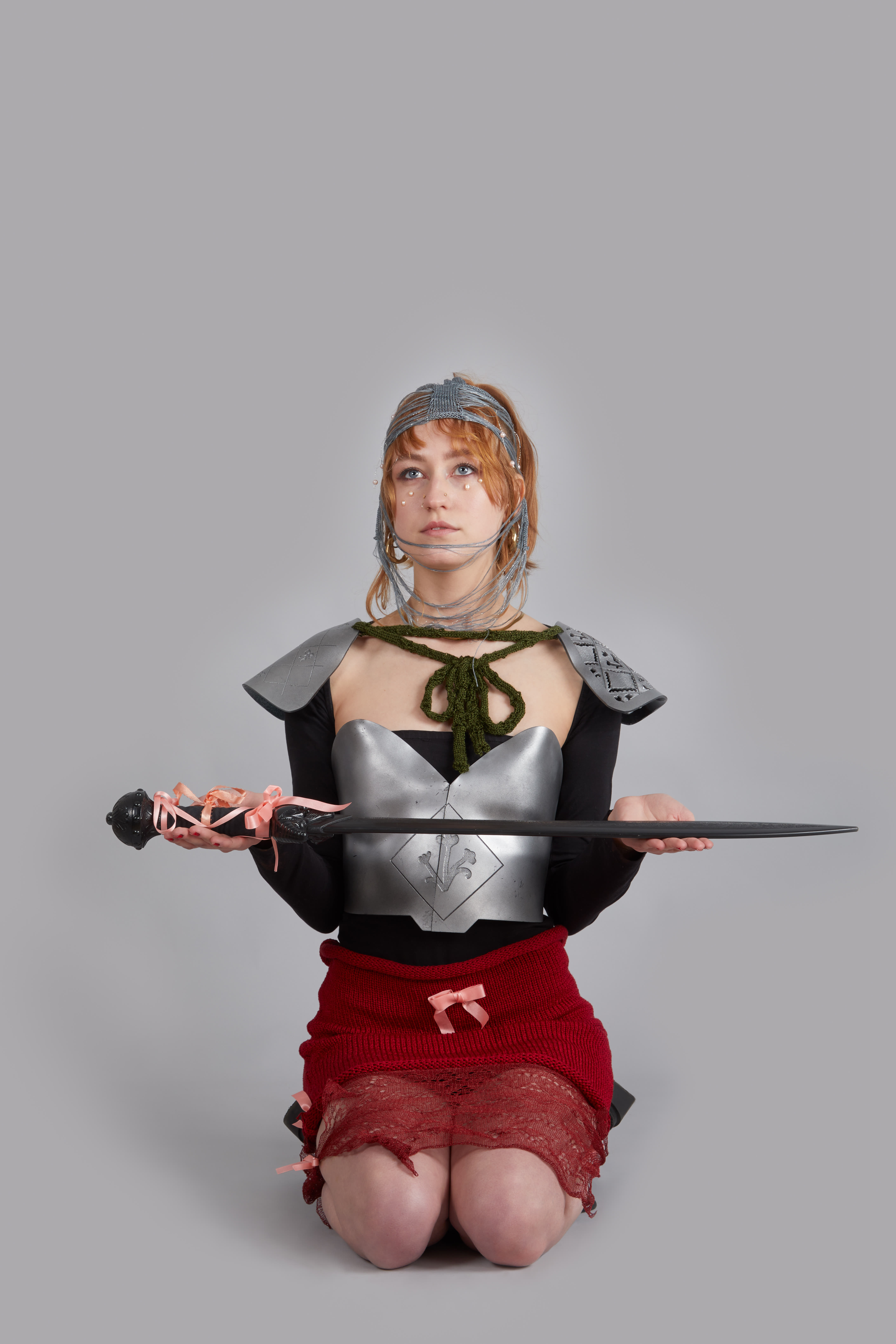 Model sat on knees holding a sword wearing armour like chest piece and shoulder pads with red knitted skirt.