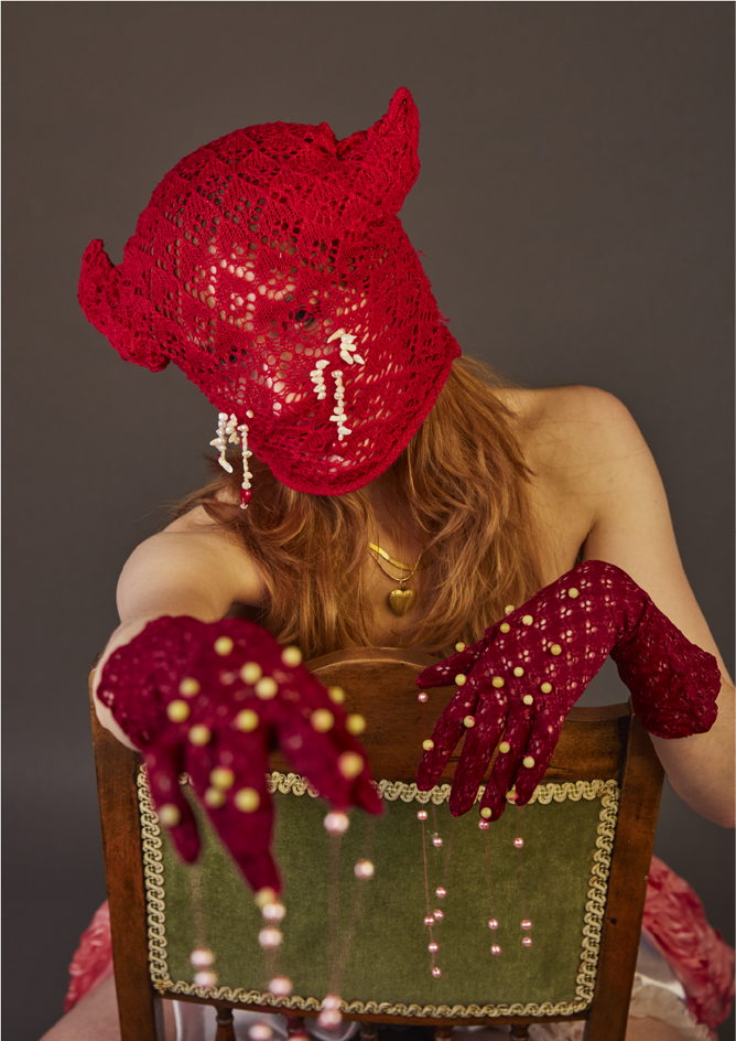 Model wearing red knit horn mask with pearls from eyes and reaching out to camera wearing maroon gloves also embellished with pearls.
