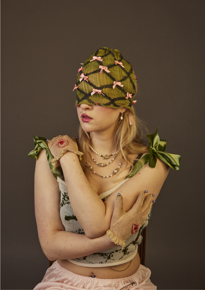 Model wearing green knit mask with pink bows covering top half of head. Models arms are wrapped round themselves wearing white and green knit top with green ribbon straps tied in bows at the shoulder.