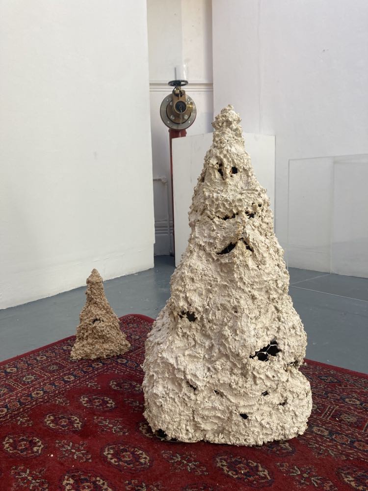 two stalagmite like forms stand on a deep red decorative persian like rug, the larger stalagmite is cream with the smaller stalgmite standing behind is rust coloured. Both works have exposed pieces of chicken wire in the form.