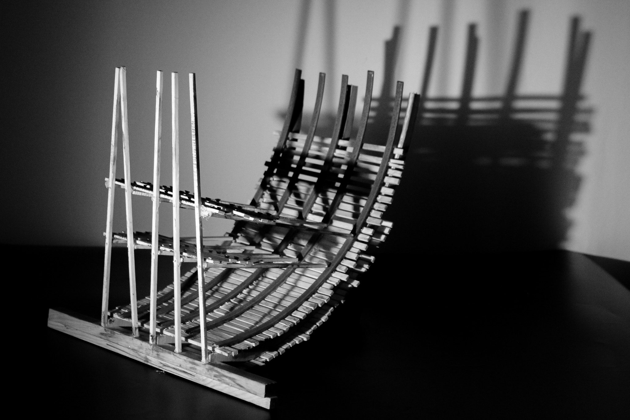 The image shows a delicate shipwreck model made from plywood and steel which sharp shadows in the background.