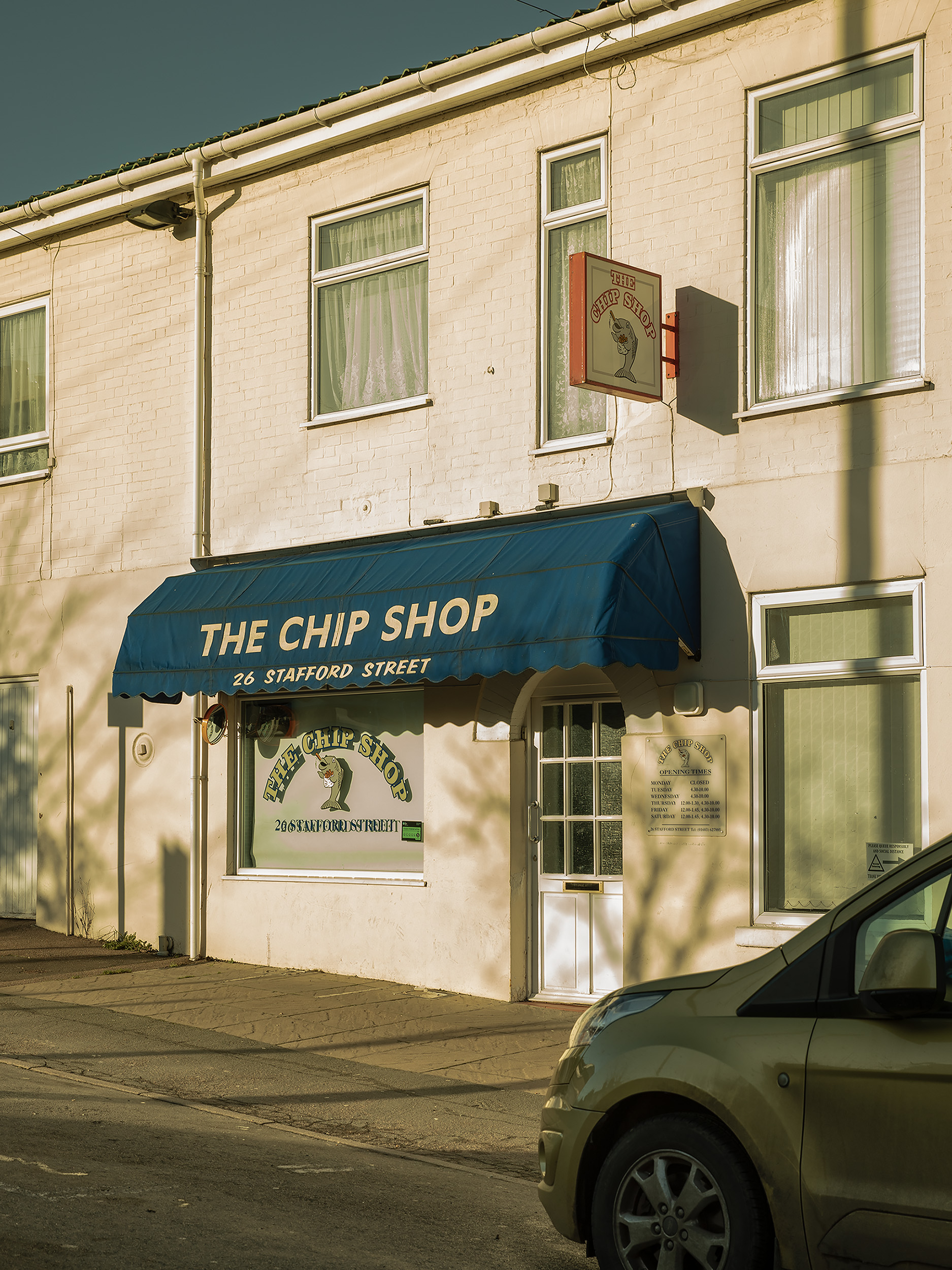 Photograph by Lauryn Boon showing a fish and chip shop with shadows of trees projected onto the walls and a car parked outside.