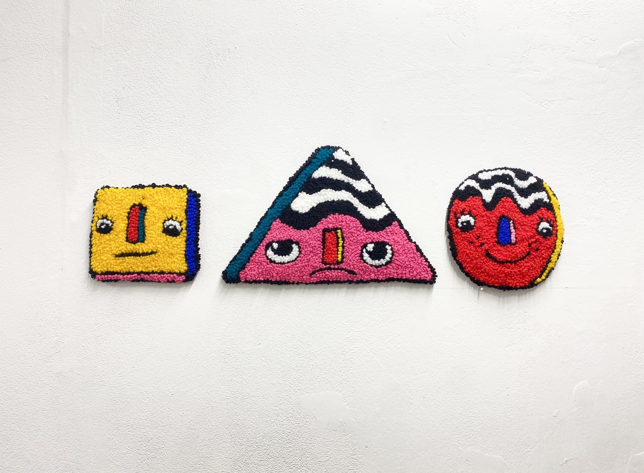 Fine Art work by Lily Bates showing a series of three brightly coloured hand tufted tapestries with faces on them
