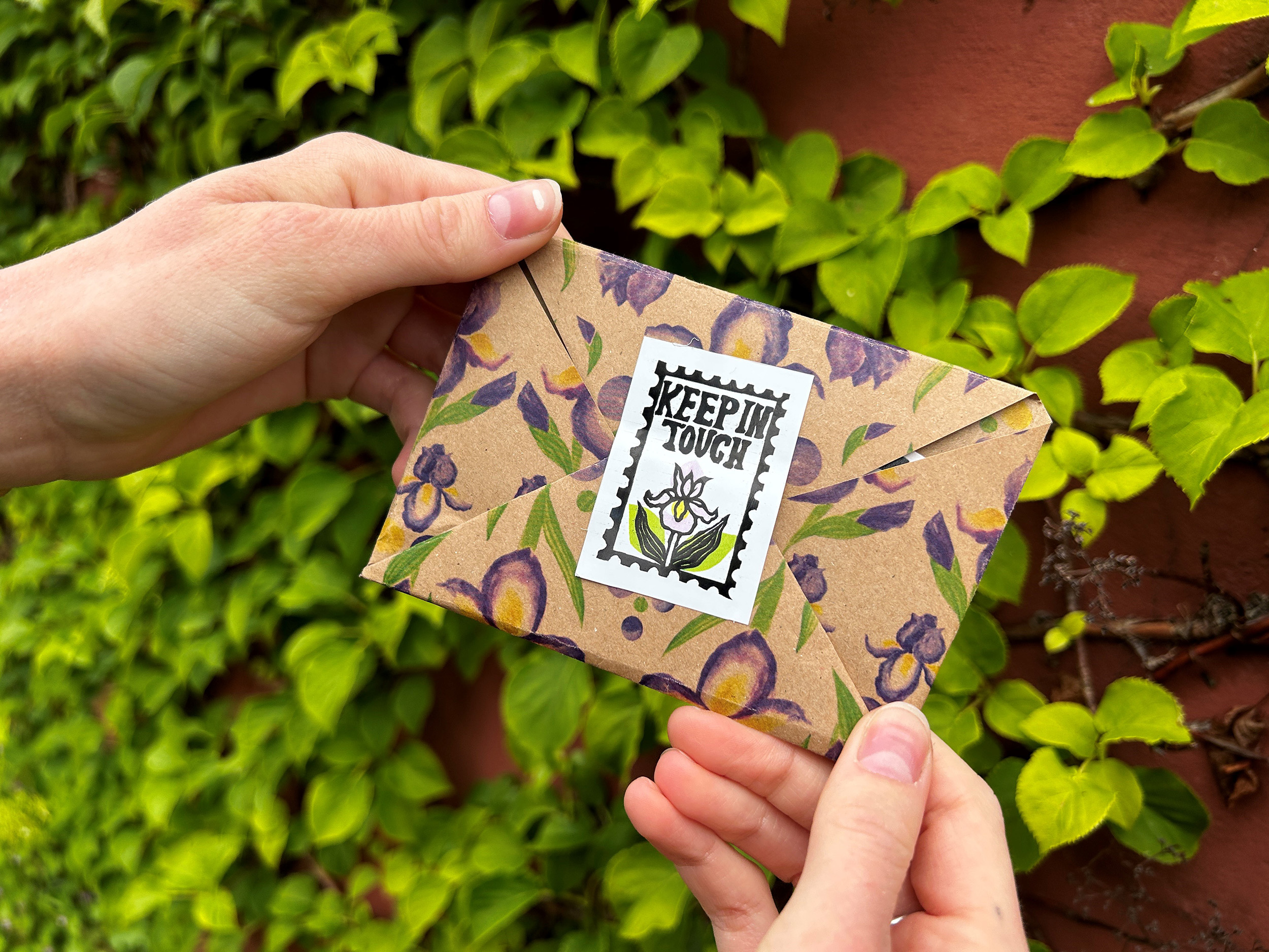 Two hands holding a decorated envelope in front of a wall. The envelope has a pattern of watercolour iris flowers and a sticker sealing it which reads 'Keep in Touch'.