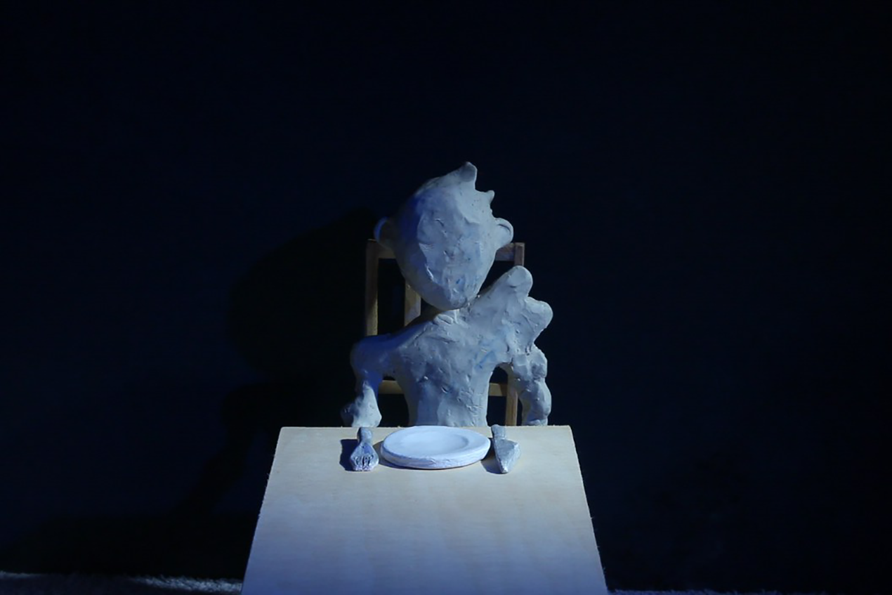A dark cold scene with a stop-motion animation puppet sitting by a dining table with and empty plate and cutlery.