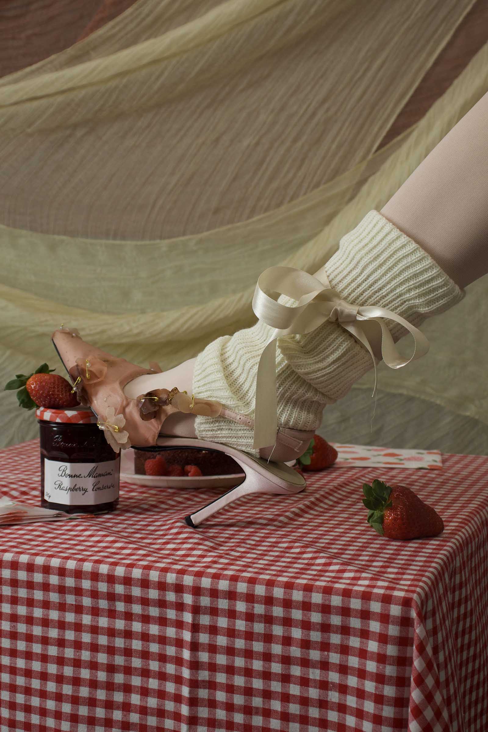BA Fashion Design work by Mollie Green showing raspberry jam jar on red gingham table with models leg wearing pink heel with cream ribbon leant againt it.