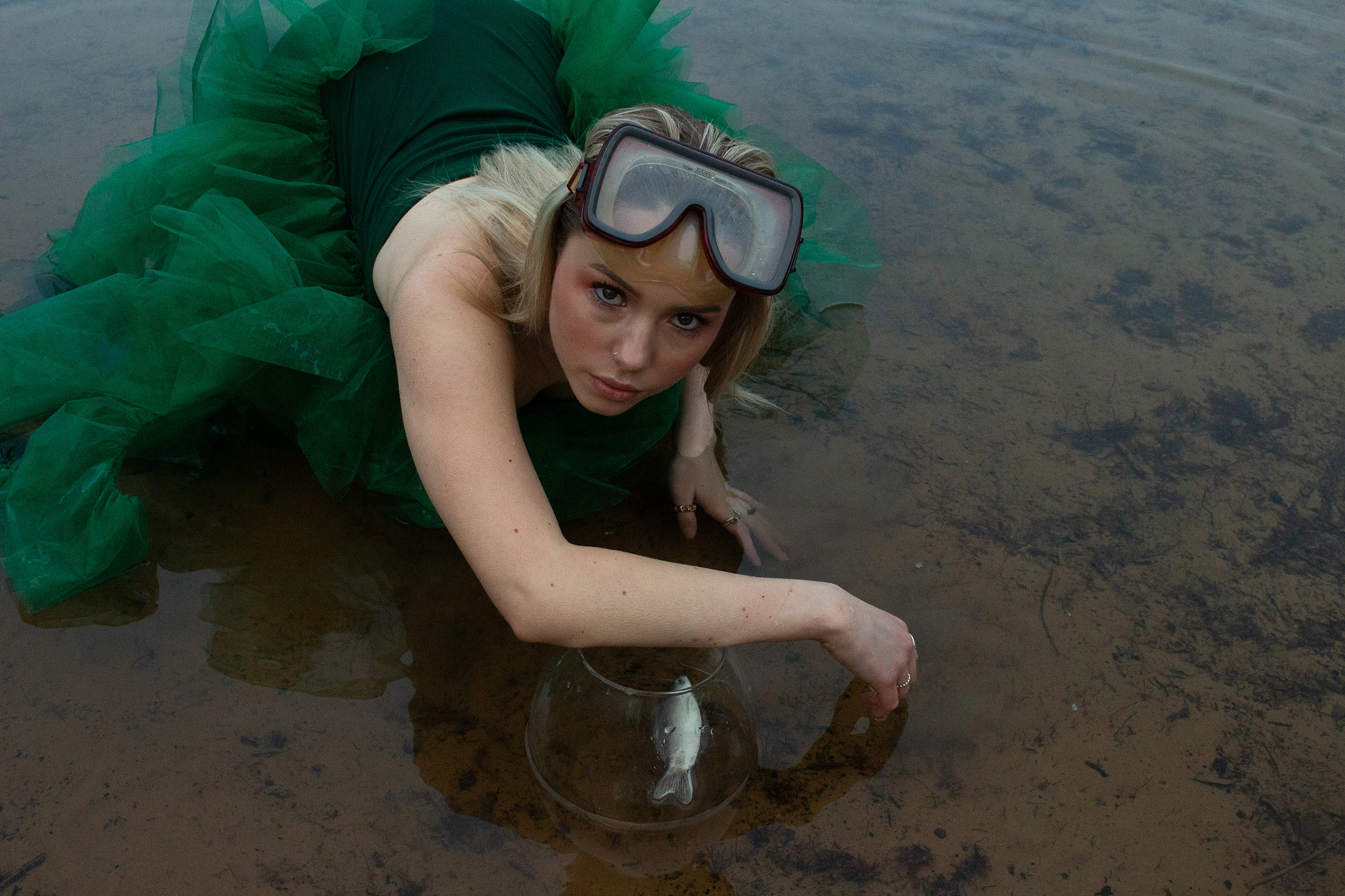 Photograph of model in green dress in shallow water with scuba goggles on and an arm over a fish bowl with a toy fish in it.
