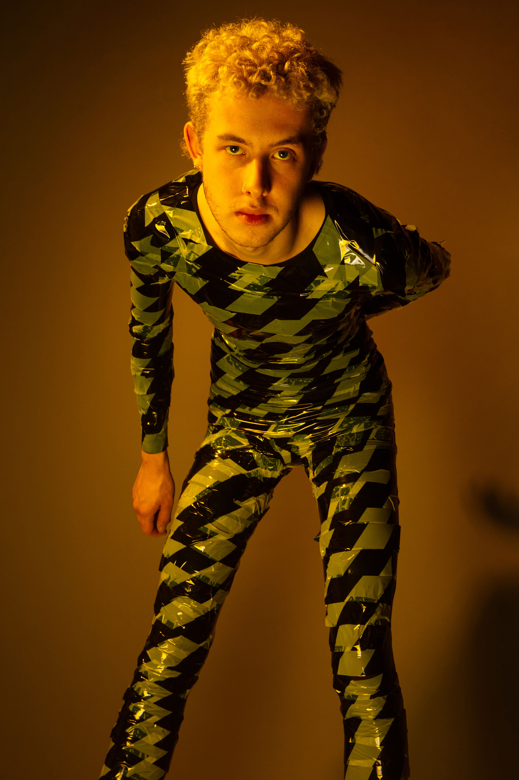 Photograph of model in plastic tape jumpsuit leaning towards camera against a yellow backdrop. Creative Direction and Styling by Rosie Parnham. Studio Photography by Ruby Williams.